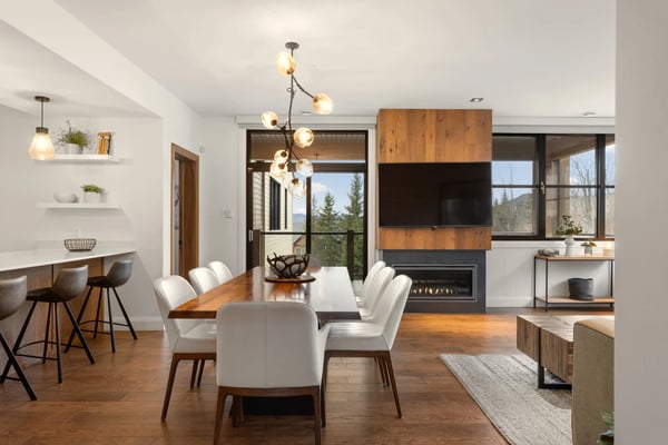 Open-plan living space with dining table, white chairs, fireplace, wood cabinetry, and mountain views through large windows