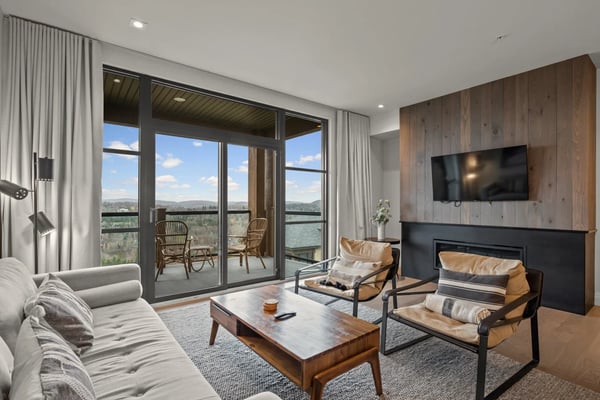 Modern living room with floor-to-ceiling windows overlooking mountains, fireplace, TV, and furnished balcony