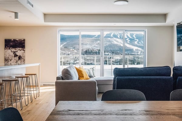 Modern living room with gray and navy seating, bar stools, and floor-to-ceiling windows overlooking snow-covered mountains and ski slopes