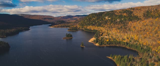 Vue aérienne d’un lac du Québec entouré d’une forêt automnale et de montagnes ondulantes sous un ciel spectaculaire
