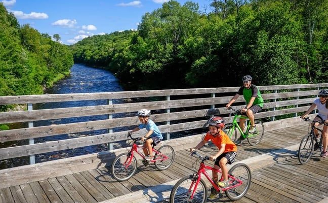 Five cyclists riding on wooden bridge over river surrounded by dense green forest and hills